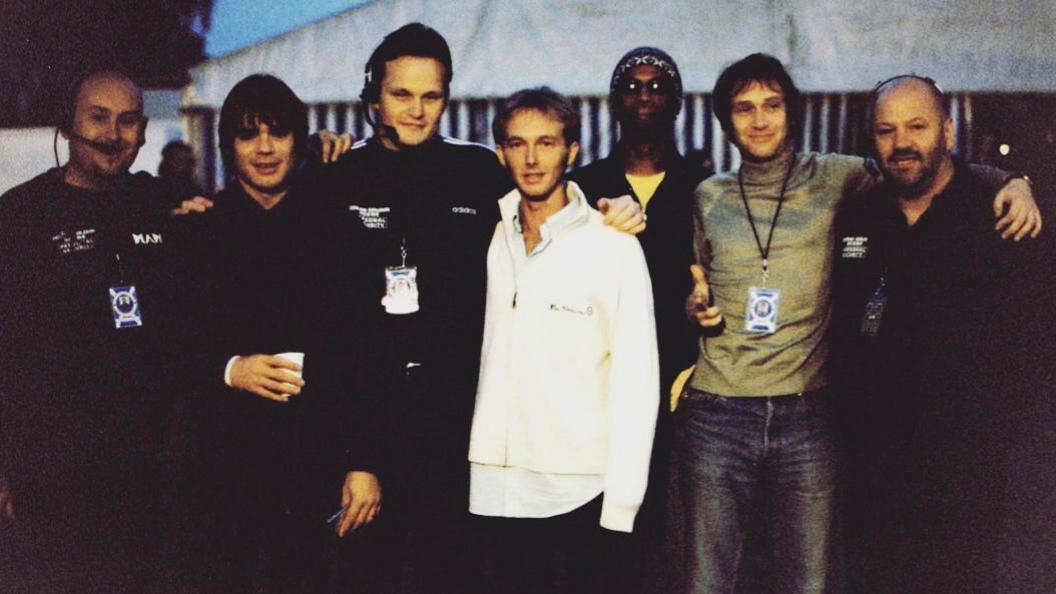 A line-up of seven men assembled for a photograph. Five have black tops, the man in the centre has a white top and the man second from right has a green top