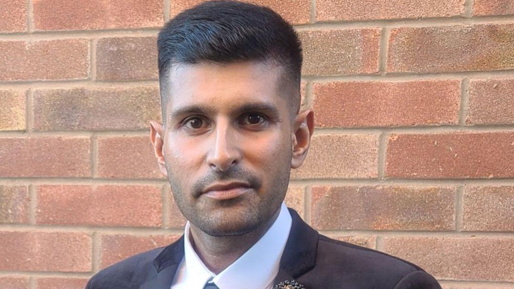 A man in a dark suit stands next to a brick wall