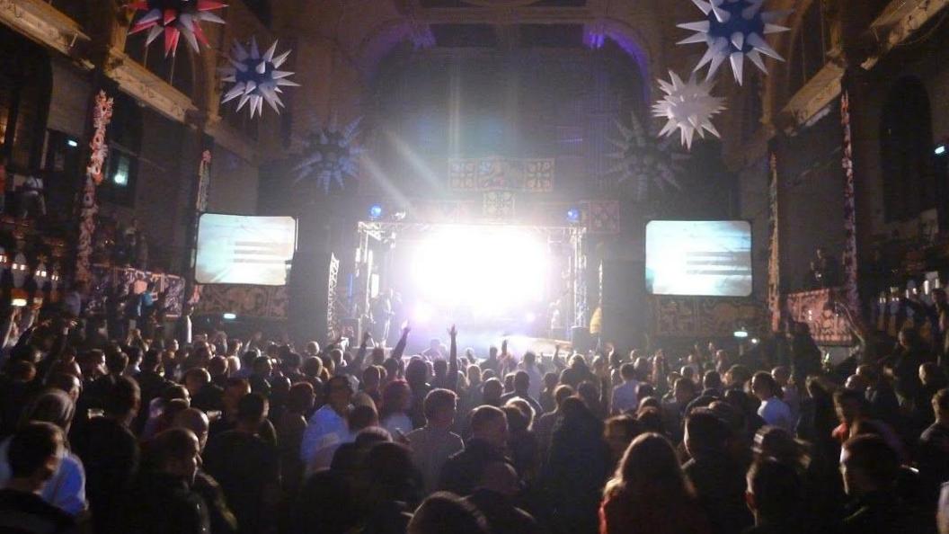 A large crowded hall with people standing in front of a stage. There are screens on either side of the stage.