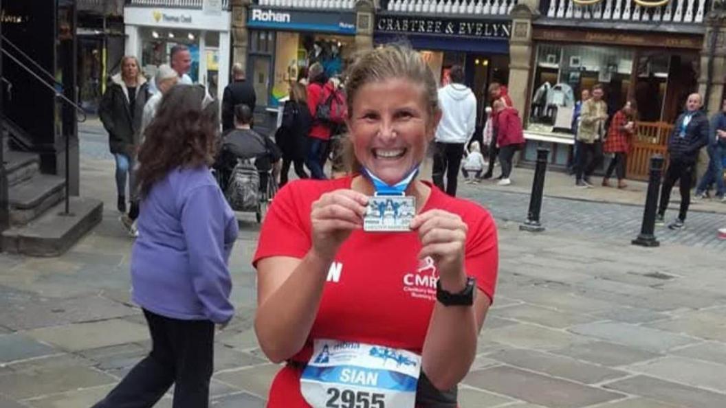 A smiling woman wearing a red sports top and running number, hold a medal round her neck. The background is a busy town centre with shops and people.