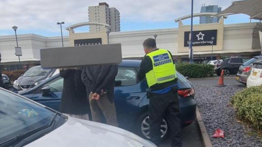 Three people next to a blue car have their backs to the camera. The signs for two stores are in the background, along with two tower blocks.