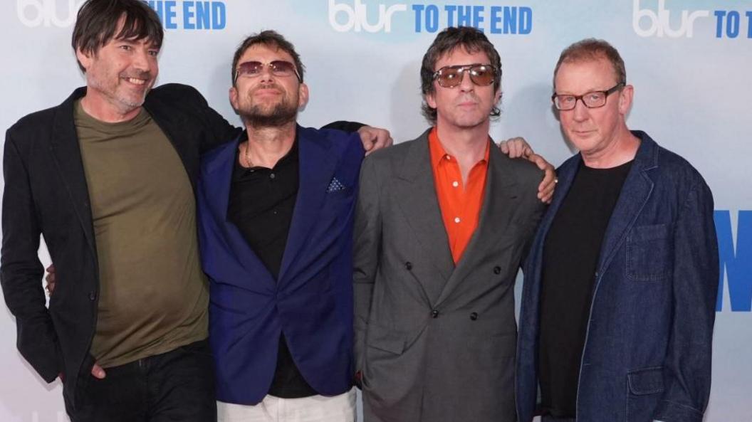 Alex James is smiling at the camera and is wearing a black suit jacket, green T-shirt and black trousers and has his arm round Damon Albarn, who is wearing a navy suit jacket, a black shirt and cream trousers as well as tinted sunglasses. He has his arm round Graham Coxon, who is wearing an orange shirt, grey suit and sunglasses. He has his arm round Dave Rowntree, who is wearing a black T-shirt and denim suit jacket.