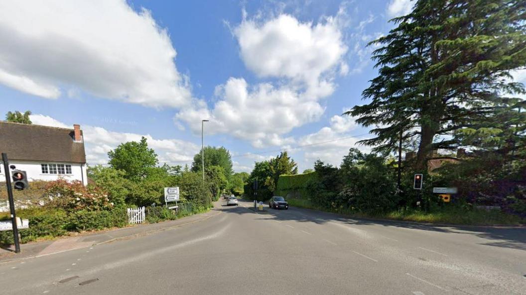 A junction with a two cars visible - one parked and one driving. There is a white building to the left and trees to the right of the road. 