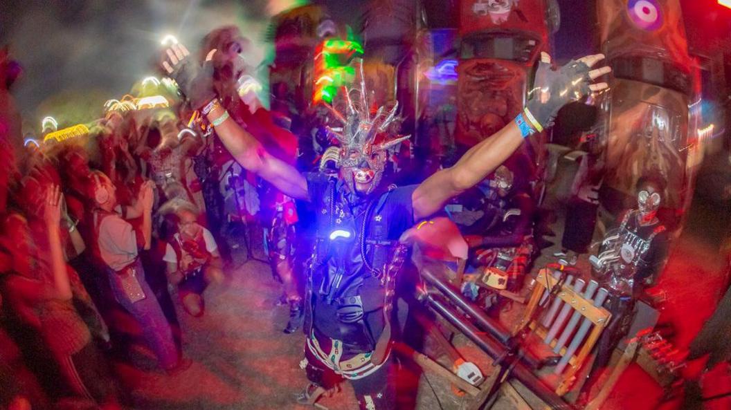 A picture shows Carhenge by night, with a performer wearing a large voice and distinct red and blue lighting