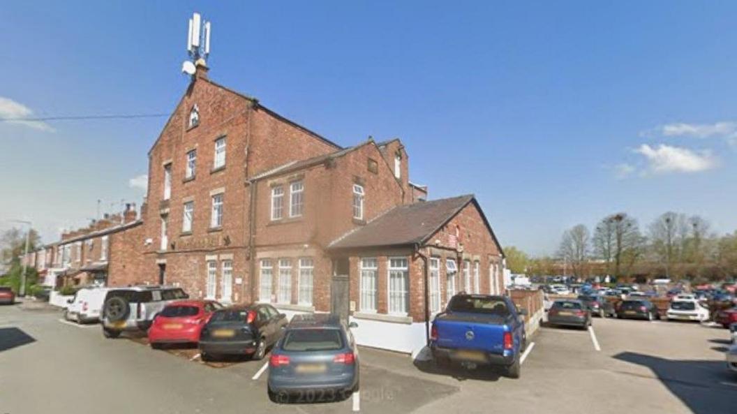 A street view of a large red brick building with cars parked outside. On the apex of the roof at the front of the building sits a large white three pronged mast.