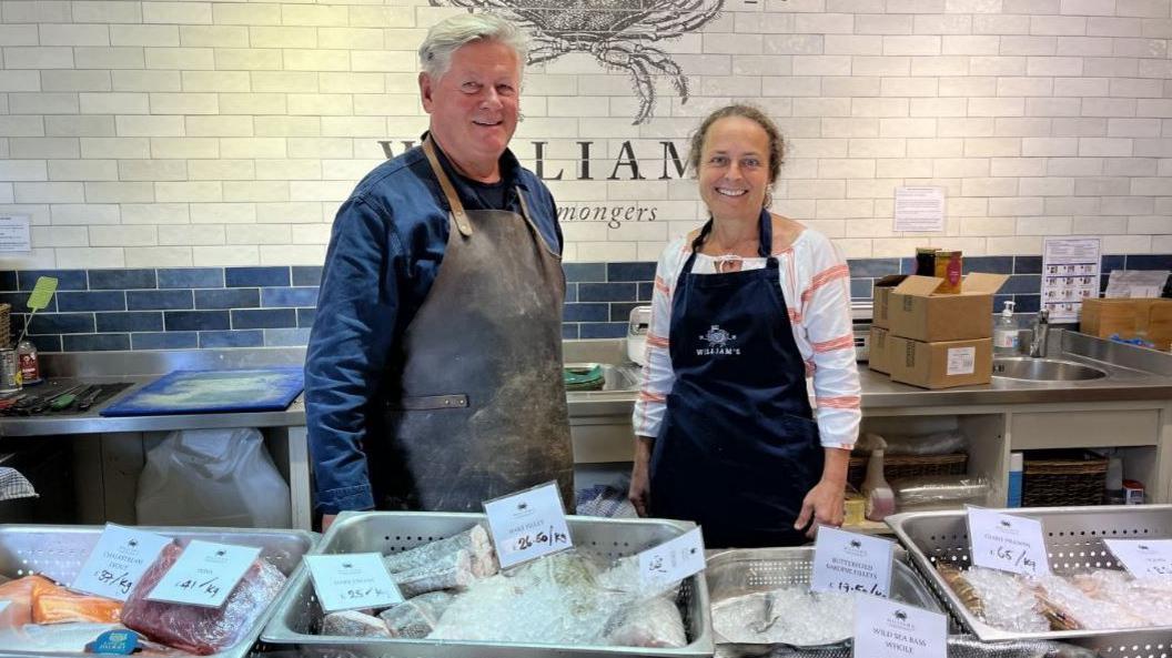 The image shows two individuals standing behind a counter at a fishmonger's stall, both wearing aprons branded with the logo "WILLIAM'S Fishmongers." In front of them, trays are neatly arranged and filled with a variety of fresh fish and seafood, each clearly labelled with prices per kilogram. The labels include: Ocean Trout – £37/kg, Tuna – £41/kg, Flathead Fillets – £26.65/kg, Hapuka Fillet – £59/kg, Giant Prawns – $65/kg, Butterflied Snapper Fillets – £49.50/kg, and Wild Sea Bass Whole – £38/kg. The setup suggests a clean, professional market environment focused on quality seafood offerings.