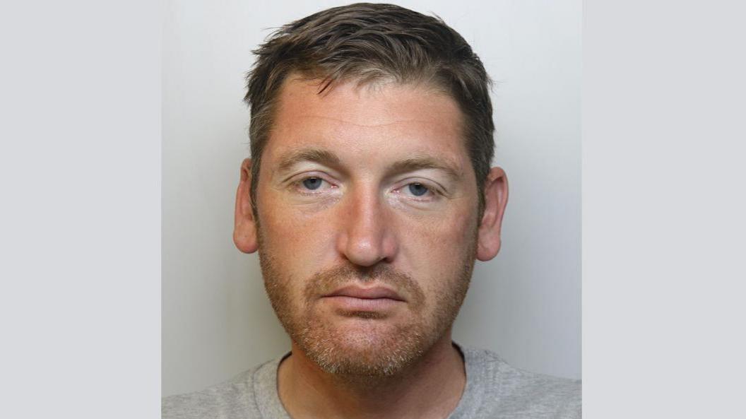 A police mugshot of Daniel Bennett. He is wearing a grey t-shirt and standing against a grey background. He has dark brown hair which is shorter on the sides, and a light brown beard. He is looking at the camera.