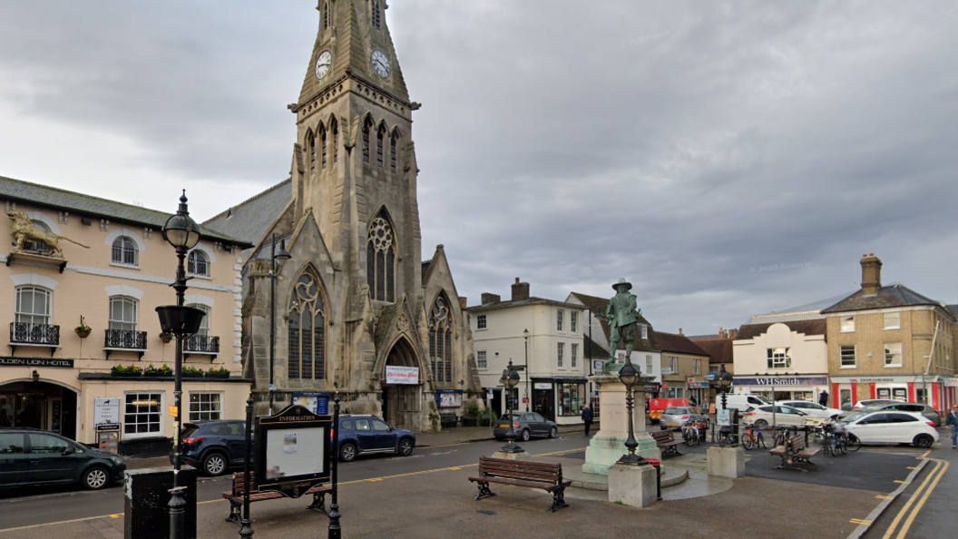St Ives' Market Hill, featuring The Free Church on the far side. In a central paved area is Oliver Cromwell's statue and street furniture. There are cars parked in front of the church and in the centre of the market place.