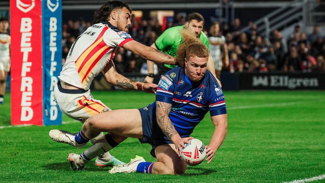 Wakefield Trinity prop Caius Faatili, in blue kit, slides on one knee to touch the ball down for a try as Catalans Dragons full-back Charlie Staines, in white with red and yellow trim, tries to grab him.