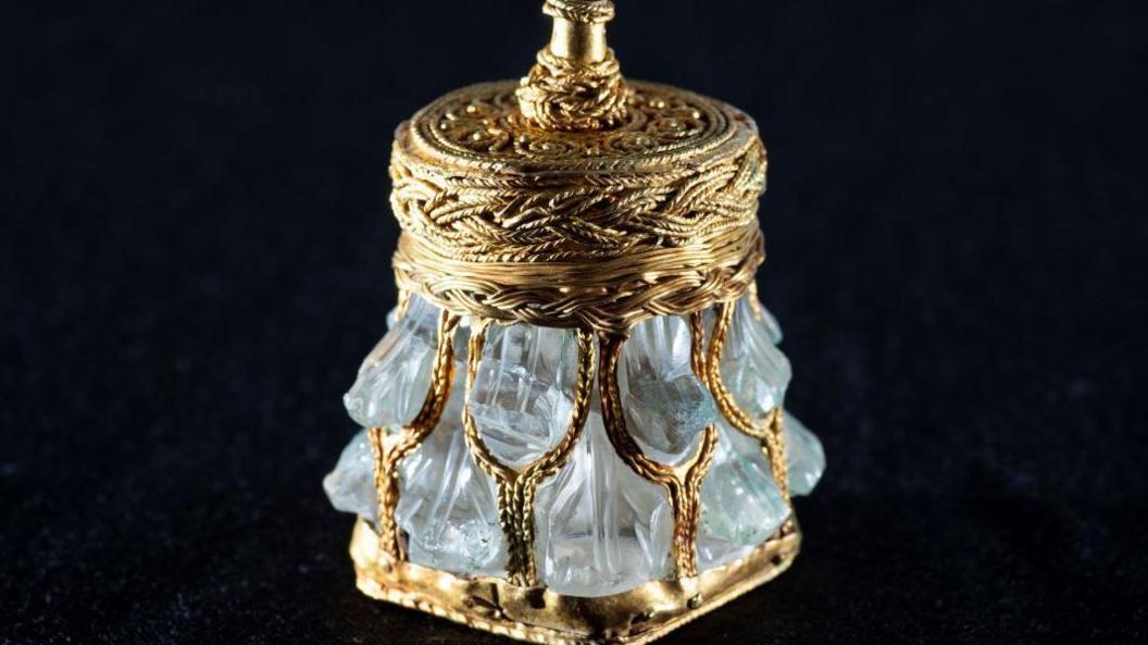 A rock crystal jar, which resembles an ornate perfume bottle decorated in gold embellishments on a black background.