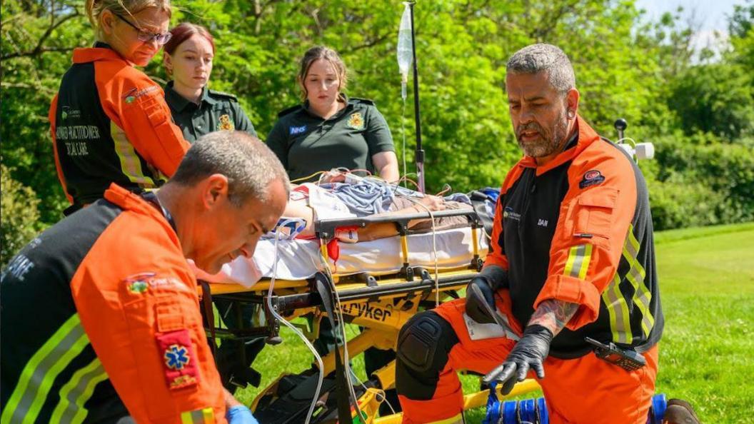 Five people dressed in green polo shirts and orange hi-vis uniforms tending to a mannequin during a simulation training exercise. They are in a field on a sunny day, surrounded by trees and grass. There is a stretcher in the background with the mannequin on, hooked up with medical tubes and covered with a blanket. Two men are kneeling in the foreground, tending to another mannequin patient on the ground.