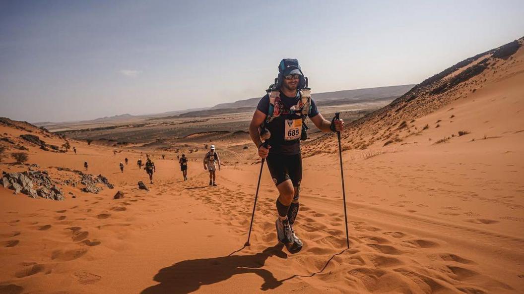 Adam, using two walking poles and carrying a large backpack, walking on some sand dunes