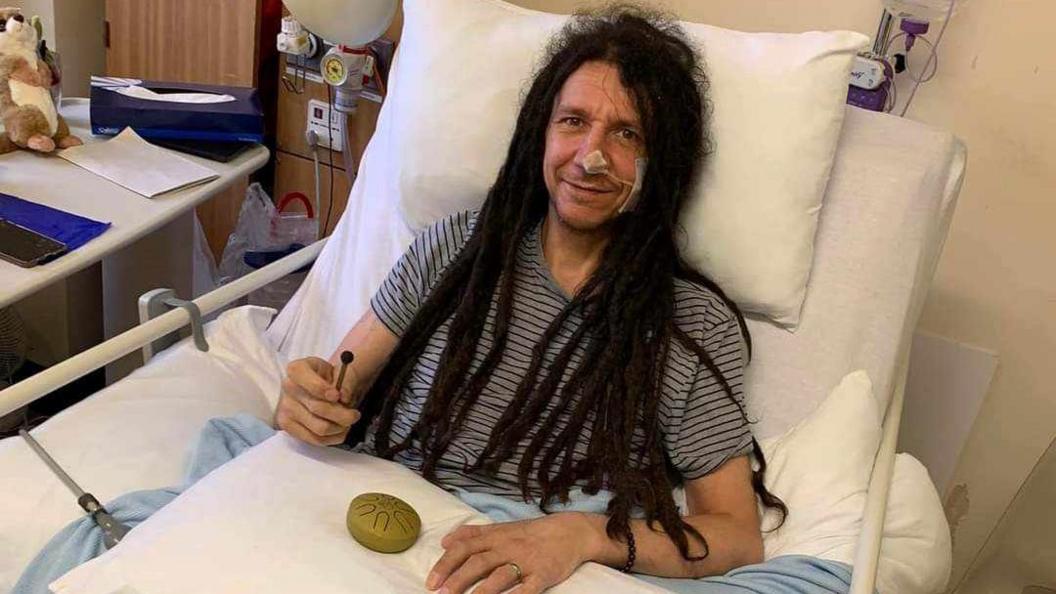 Tony sitting in his hospital bed. He has a grey-T-shirt on, is propped up against the pillows in his bed, has medical and is smiling towards the camera. 