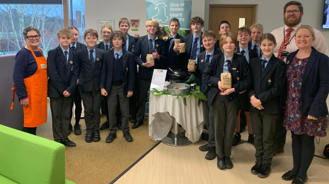 Pupils from Haberdashers’ Monmouth School and Monmouth Comprehensive, at County Hall in Usk, following a presentation urging councillors to do more to remove goods associated with deforestation from school meals. The pupil asr wearing their school uniforms, consisting of grey trousers, a blue shirt, blue tie, and a blue blazer. They are stood together, some holding little brown bags and all looking at the camera. There are also three adults (teachers) in the photo, who are stood on either end, the one on the left has an orange apron over a navy top, and smiles. The two on the right, there is a male teacher with a red tie and pink shirt, and in front of him is a female teachers with a navy blazer with a patterned dress under it.