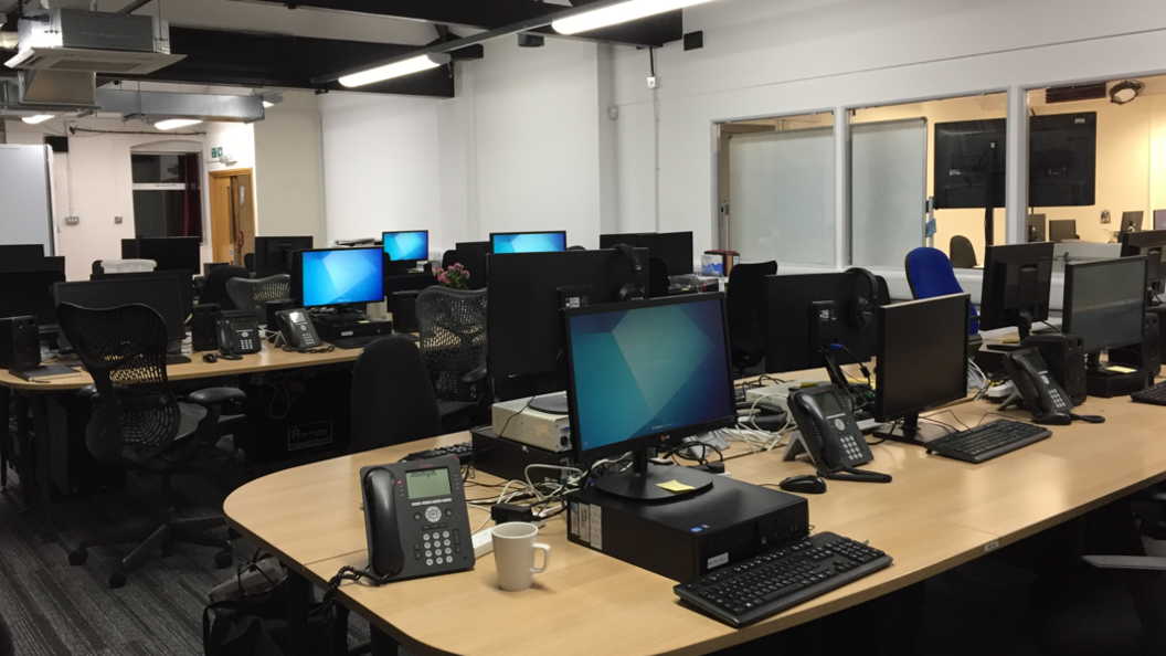 Two rows of light wood desks in an office, with a number of computers and telephones on each desk.