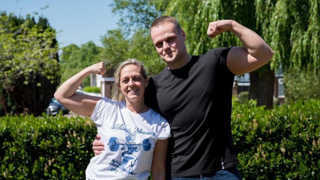 Ashli Wise with long blonde hair tied back and wearing a white T-shirt with a blue drawing of someone lifting weights, and Cory Wise who is taller than Ashli and has very short dark hair. He is wearing a dark T-shirt and jeans. They are both raising a fist in a muscle pose. They are standing in front of hedges and trees.