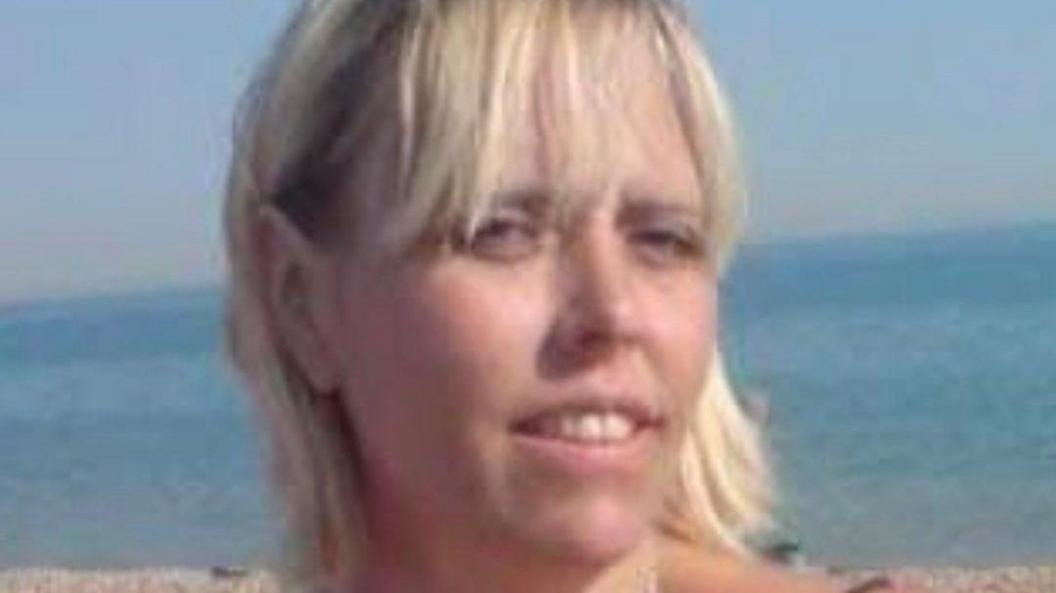 A woman with blonde hair looking into the camera. Behind her is the sea