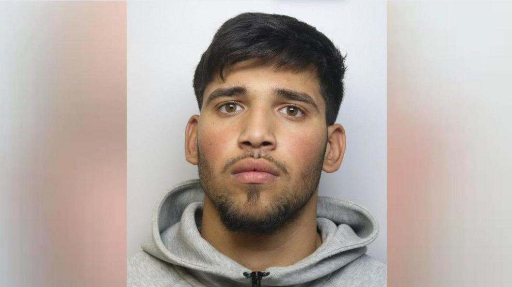 Alex Tiuga is staring at the camera. He has short black hair and a short trimmed beard and moustache and is wearing a grey hoody