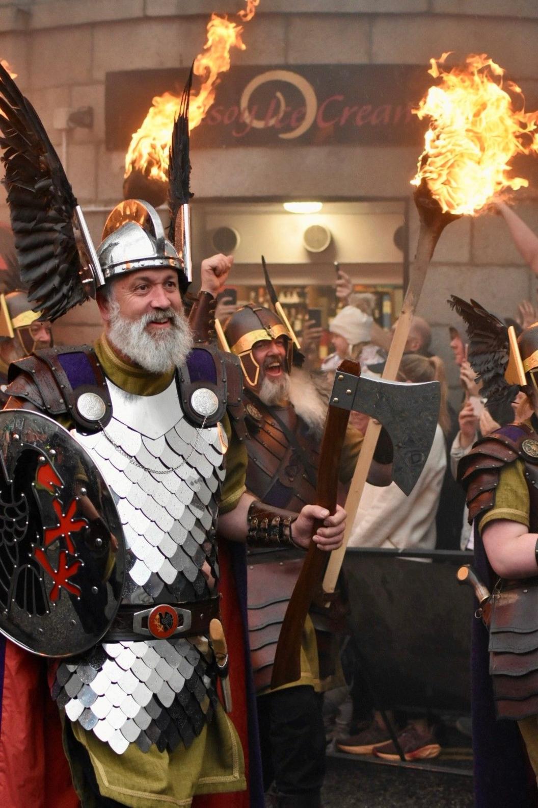 A man with a grey beard in a full Viking outfit is holding an axe and shield. There are other people in the background also in Viking outfits an with burning torches.