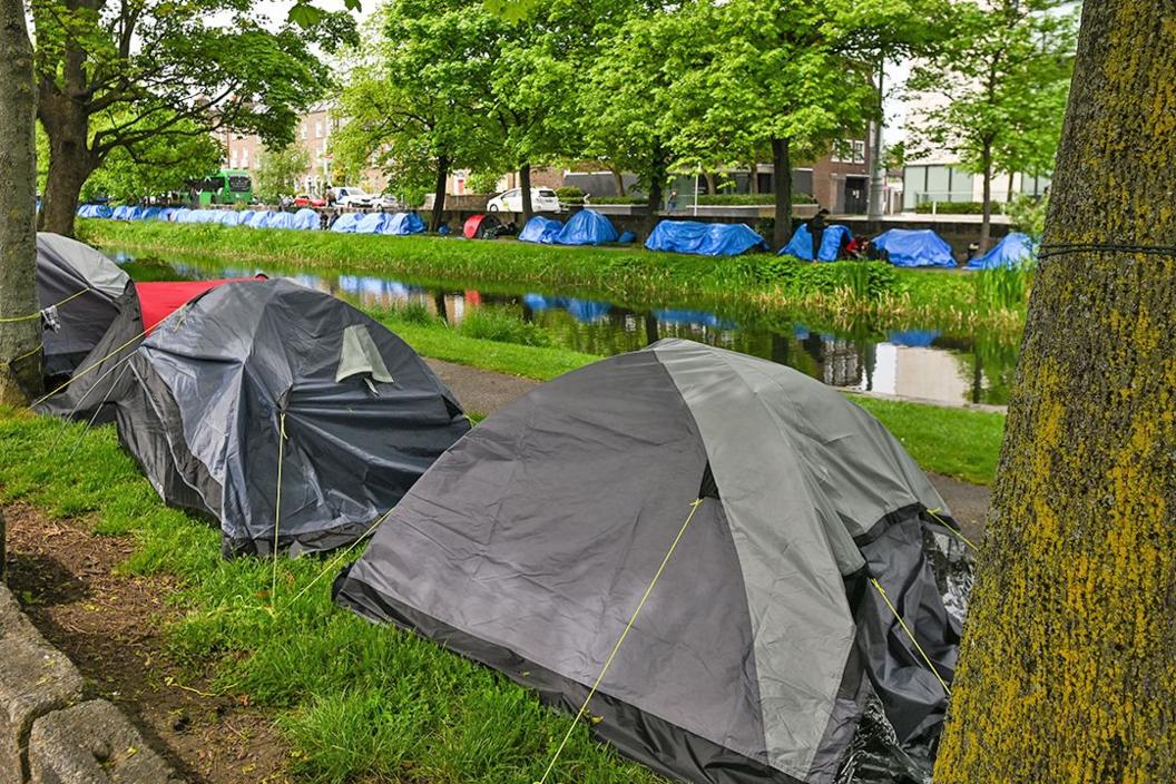 Two rows of tents - one grey, one blue - are pitched along both sides of a canal, under trees. 