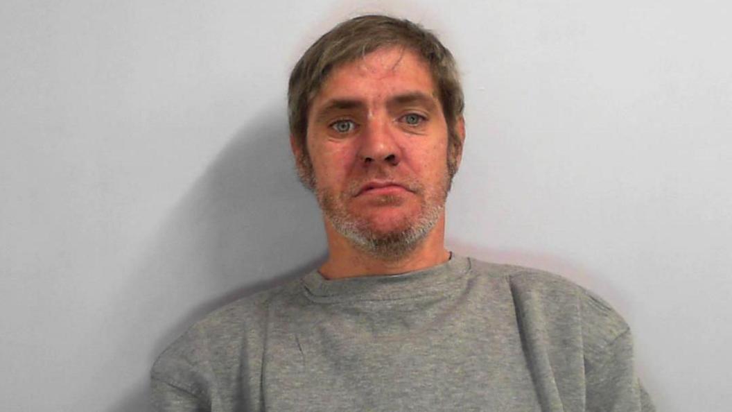 A mugshot of a man in his 40s with grey mid-length hair and grey stubble. He has blue eyes.