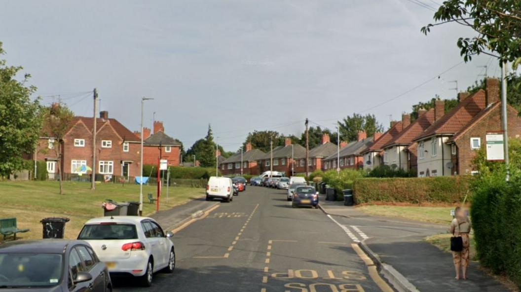 A residential street with cars parked on either side. There are yellow bus stop markings on both sides of the street. On the right there is a junction leading off to another street. Red brick houses line the right hand side of the street.