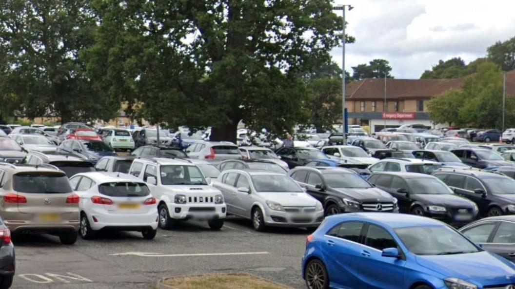 A full car park with rows of cars and a tree standing in the middle with large beige-brick buildings in the distance.