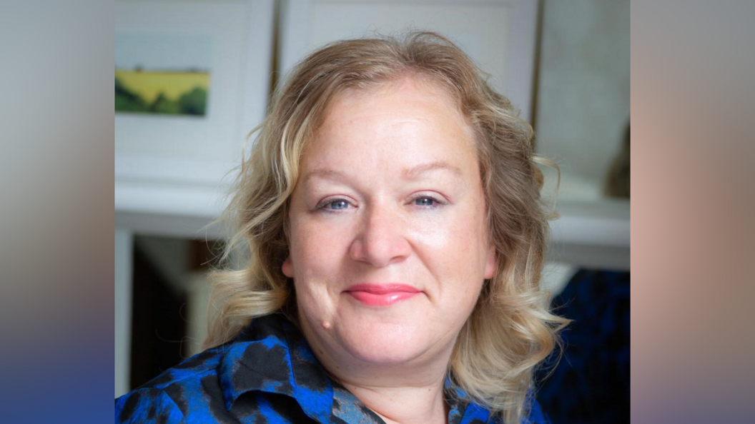 Jo Reeder smiles at the camera. She has curled, shoulder length blonde hair and is wearing pink lipstick. She wears a blue and black dress.