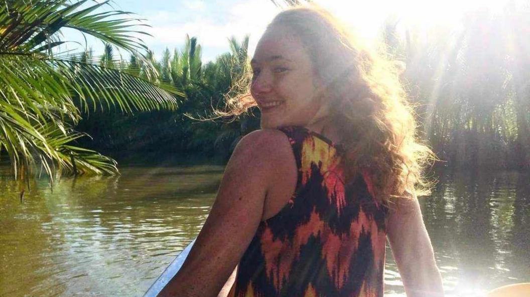 A young woman is standing with her back angled towards the camera and she is looking over her shoulder and smiling. She has wavy hair and the sun is shining down on her. She's on a boat out in a green river surrounded by trees. She's wearing a top with a black, yellow and orange design on it.