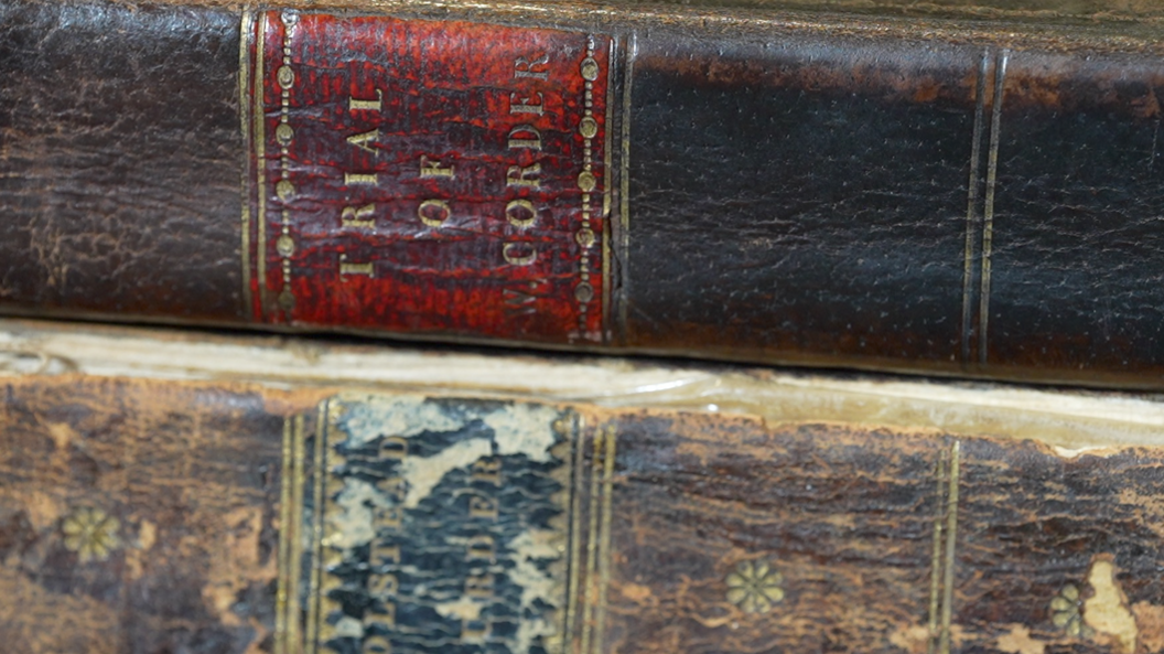 A close up of the spines of the two old, brown books. The books are on top of each other. The title of the spine of the book on top reads "Trial of W Corden". The title on the book underneath reads "Polstead - William Corder".