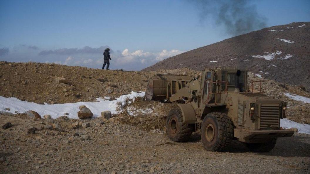 An Israeli soldier stands on top of a rocky hill, some snow seen on the ground, with a dirt-coloured bulldozer below the person 