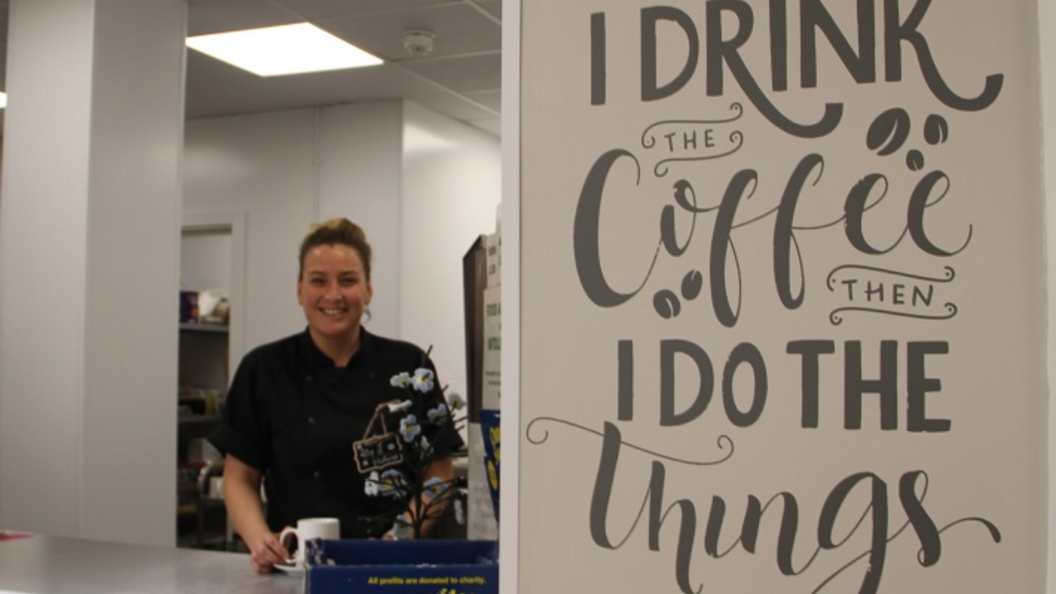 A smiling cafe worker standing behind a table top. She has blond hair which is in a bun and a black chefs coat. There is feature wall that says "I drink the coffee then I do the things"