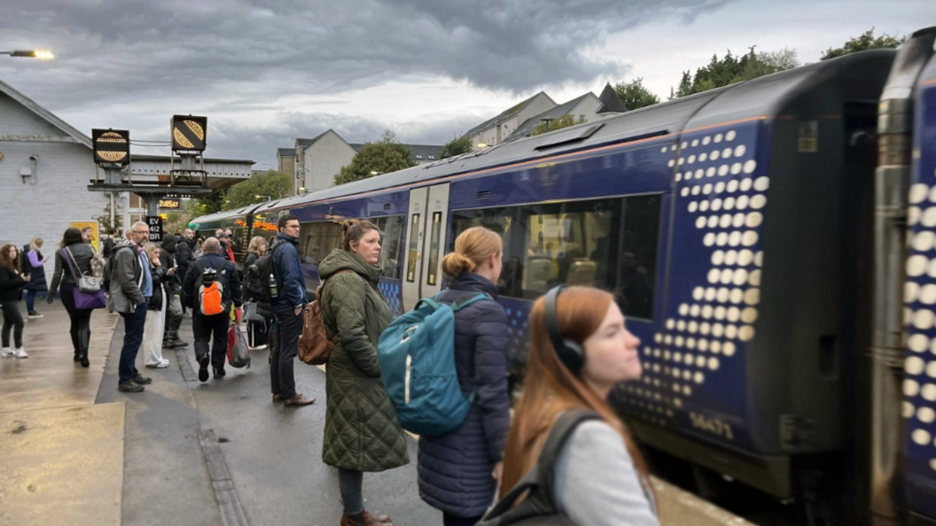 Crowd of train passengers stand on platform with blue scotrail train at station under a grey cloudy sky.