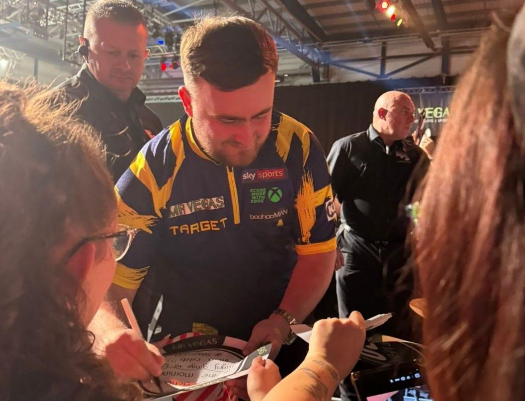 Luke Littler is in the middle of the photo, writing on something with a pen in his right hand. Two people on the far left and right of the image have their backs to the camera. Two men wearing black are either side of Littler in the photo.