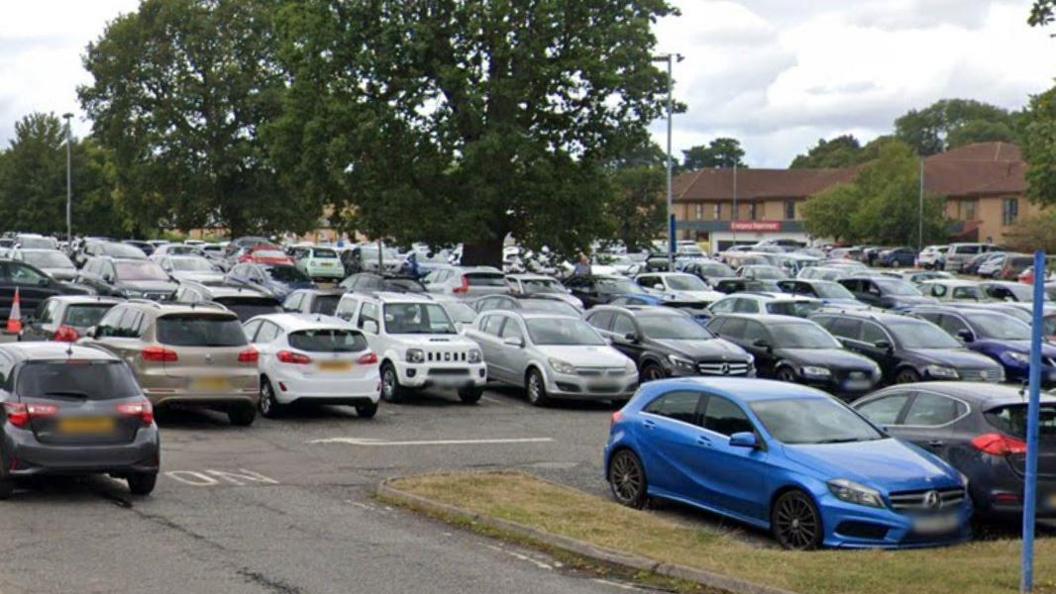 Lots of cars in a crowded car park with some trees and light brown buildings in the background