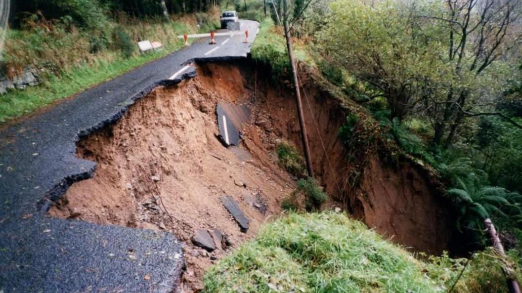 An earlier landslide on the Glenshesk Road which happened in March this year.   A large section of the road and its verge have fallen several feet down a hill, leaving a steep cliff edge. Sections of broken tarmac are lying on top of the exposed soil.  The areas is cordoned off with traffic cones and tape. 