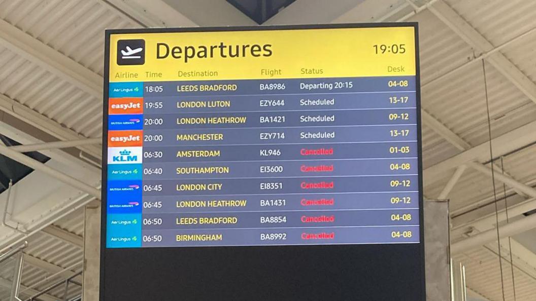 A departures board at Belfast City Airport. It lists flights from Leeds Bradford, London Luton, London Heathrow, Manchester, Amsterdam, Southampton, London City, London Heathrow, Leeds Bradford and Birmingham. Beside the six final flights is cancelled in red font.