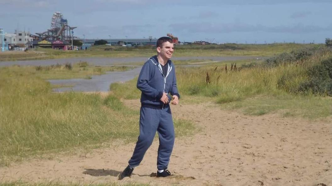 Declan running about near the seafront in Skegness. He is wearing a blue tracksuit and black trainers. A Ferris wheel and funfair in the background