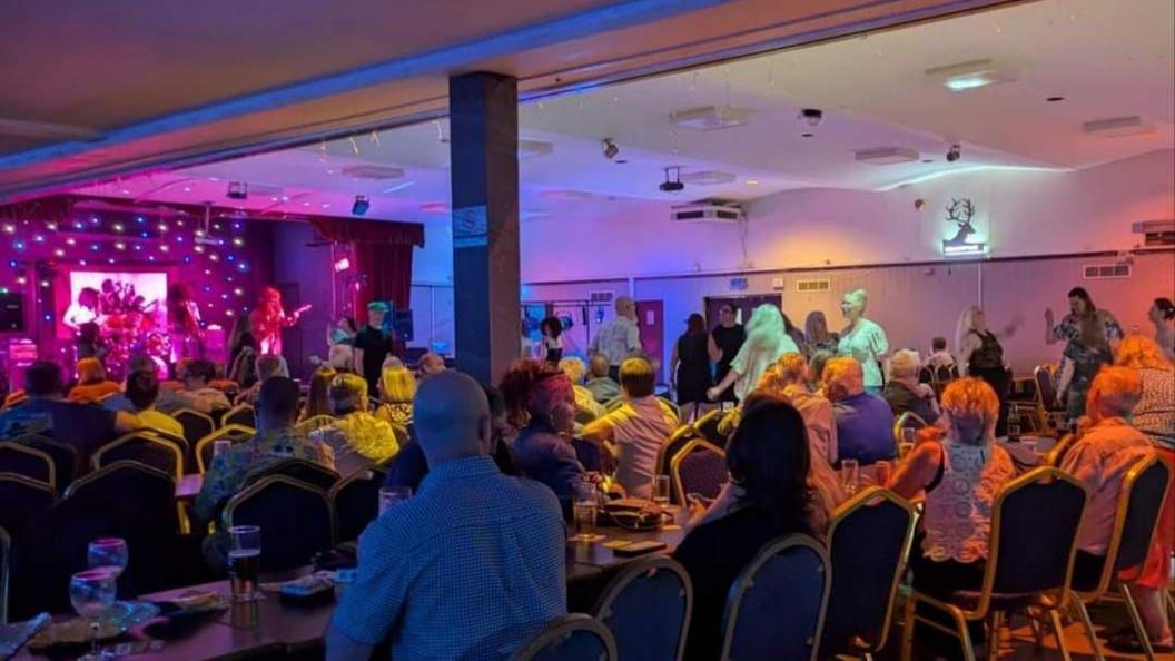 A busy function room full of people sat at tables watching a live band on stage. 