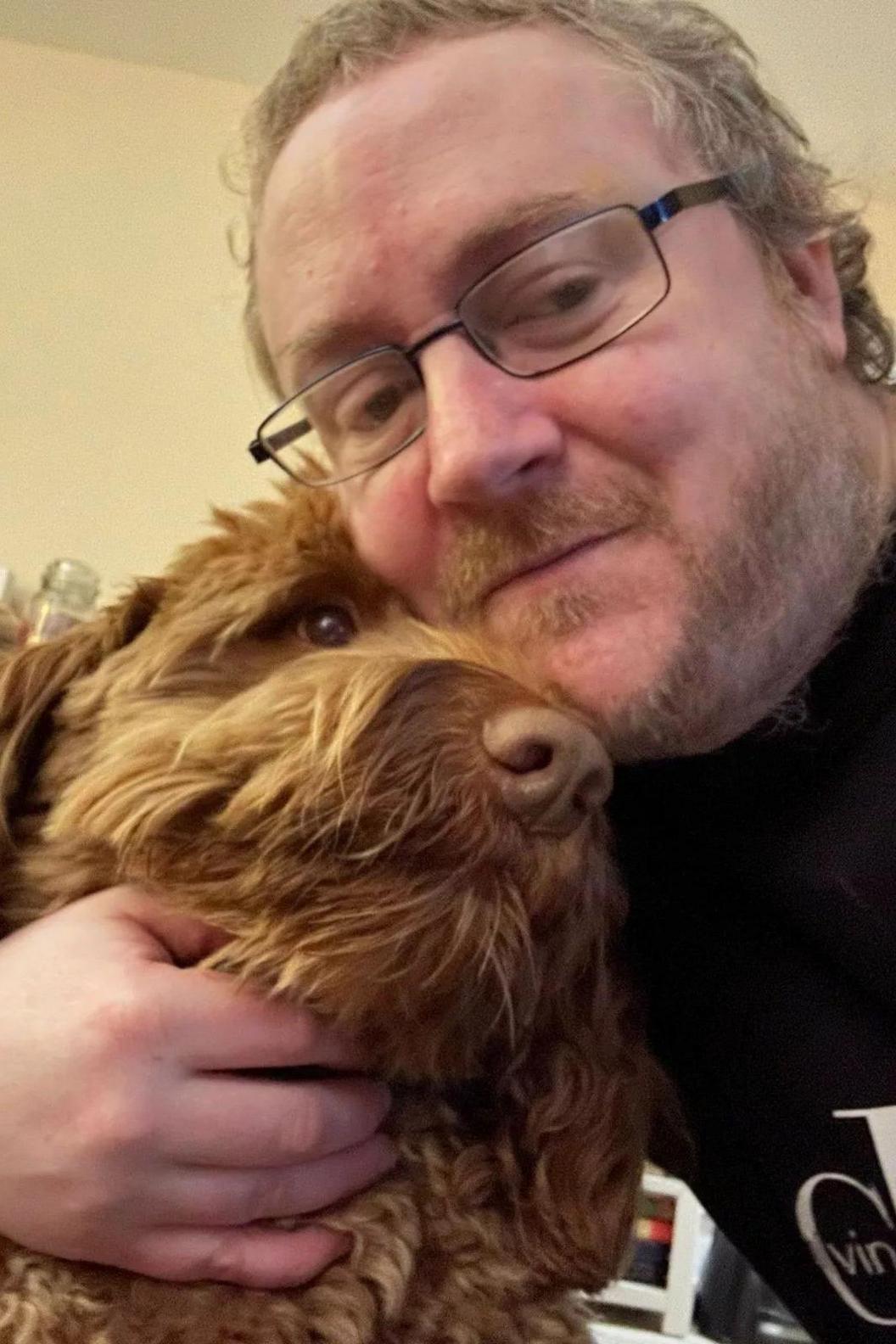 Matt Saunders is smiling at the camera as he hugs a brown Cockapoo-type brown dog.