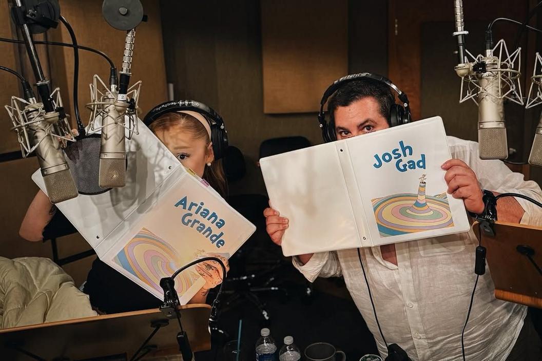 ariana grande and josh gad hide behind folders with their names written on them while sitting in a recording studio