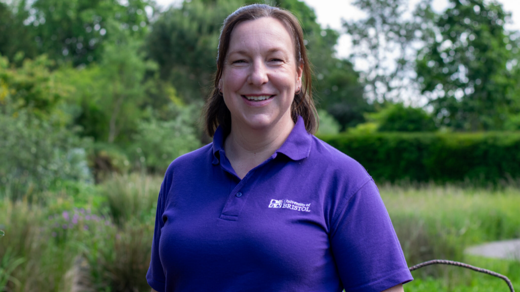 Caroline Rudge smiles into the camera wearing a purple top with the University of Bristol on it, with a garden backdrop behind her