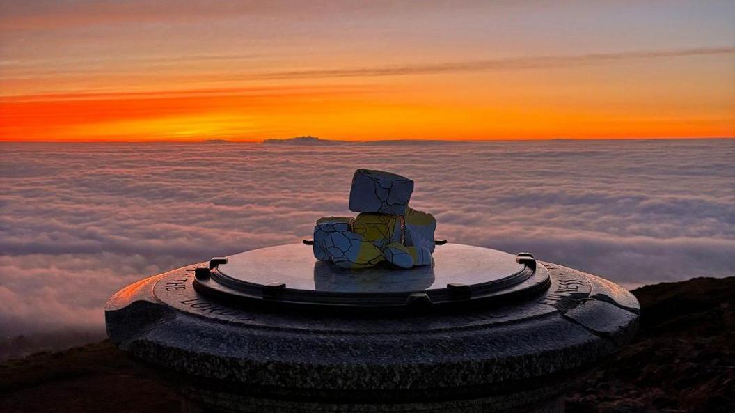 Red and yellow stripes across the sky above white clouds which cover the ground in front of a hill. The foreground shows the top of a metal circular marker at the top of the hill.