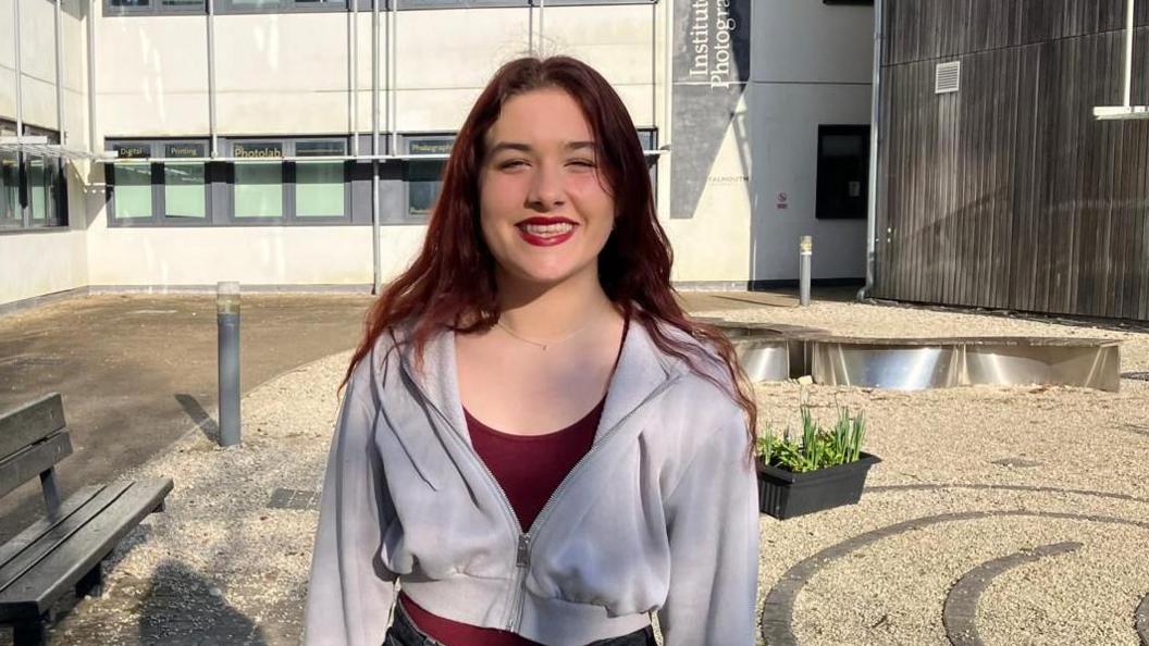 A young woman with red hair smiling, searing a grey jumper in front of a university building