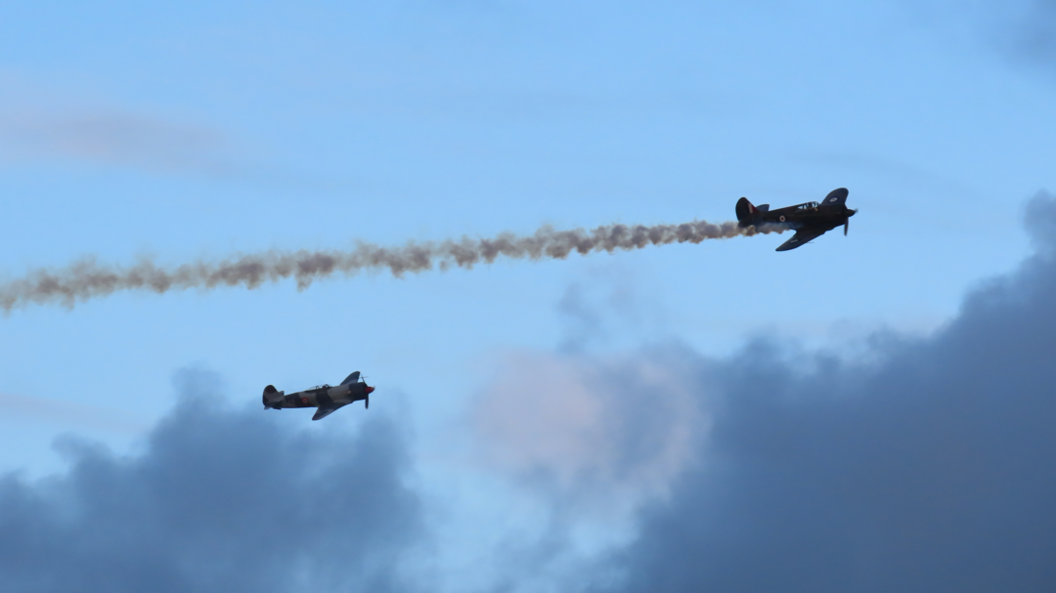 A Boomerang plane and a Yak-3 plane flying in the sky. There is dark grey smoke coming out of the back of one of them.