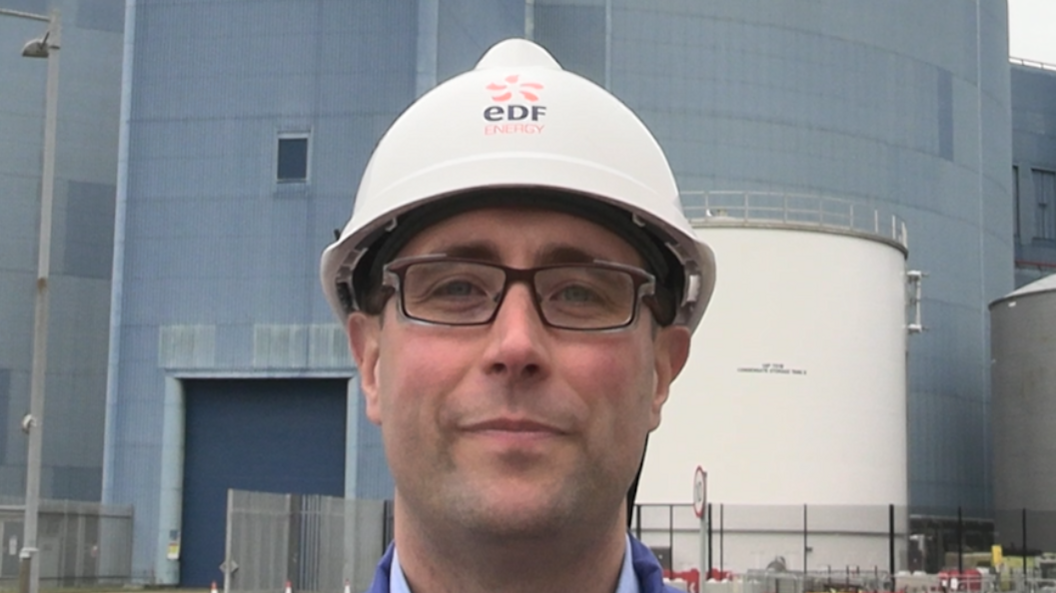 Christopher Rees smiles at the camera while wearing a white hard helmet. He is wearing glasses and a blue shirt. Behind him is the Sizewell nuclear station building which sits behind a gated fence.