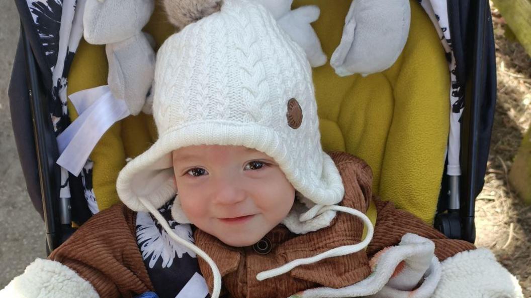 A young boy with brown eyes and wearing a white hat and brown coat smiles at the camera while sitting in a push chair