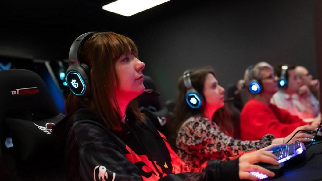 A row of people sat in chairs with headsets on looking at screens