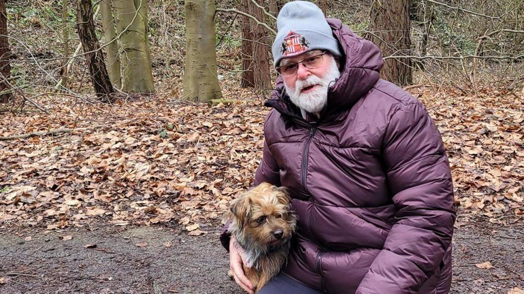 Peter Sixsmith is sitting on a bench and holding a small dog. Peter is wearing a purple puffer jacket with a hood and grey beanie with Sunderland AFC's crest on it. He has white beard and glasses. The path behind him is covered in autumn leaves.