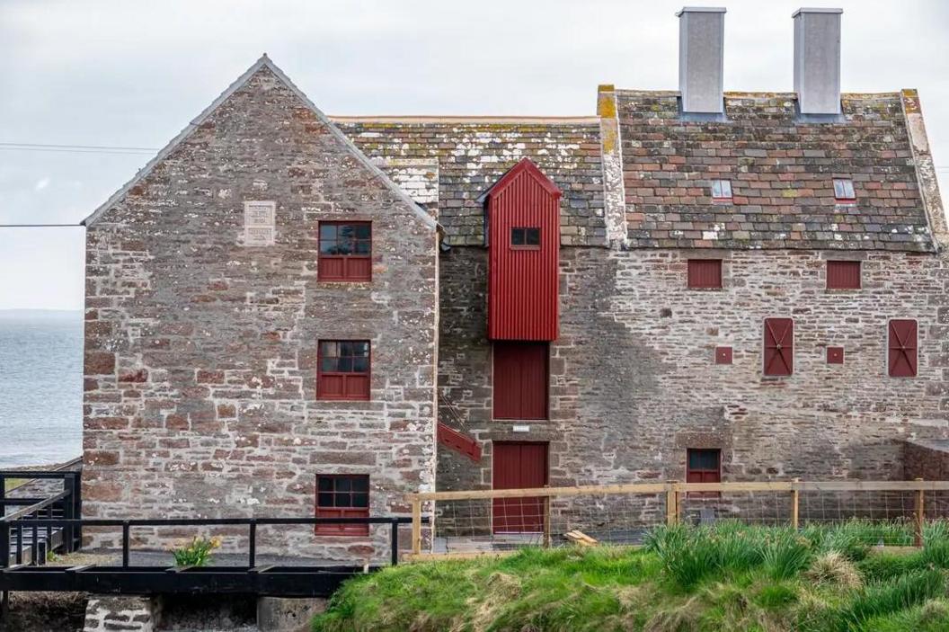 Large stone three storey building with rusty red coloured window frames and shutters. There is a walkway with railings around the outside of the building